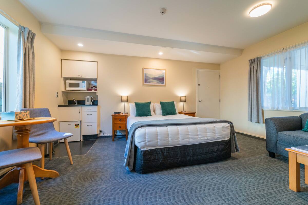 A hotel room with a bed, a desk, a chair, a sofa, a microwave, a kettle, and a painting on the wall.
