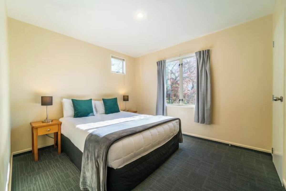 A hotel room with a bed, a nightstand, a window with curtains, and a small table lamp.