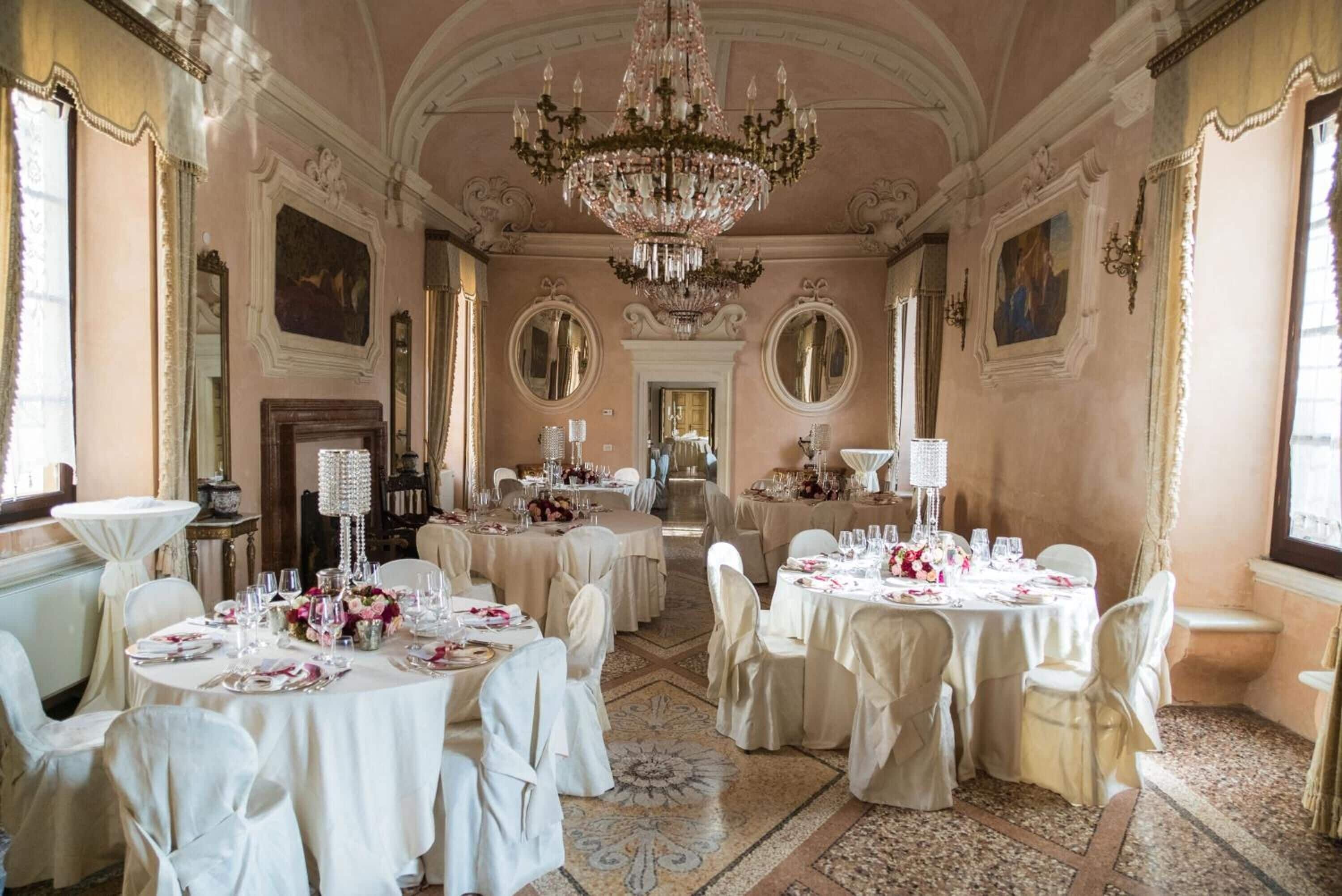 A grand dining hall with ornate chandeliers, round tables set for a formal event, and classical architecture.