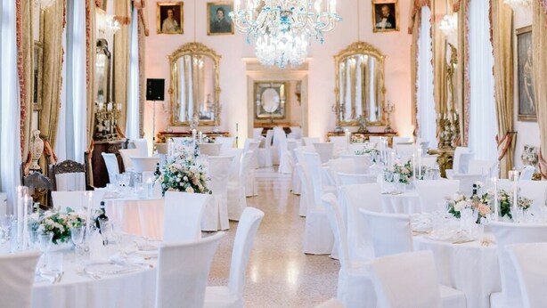 Un salón imponente con candelabros ornamentados, paredes espejadas y filas de mesas redondas preparadas para un evento formal.