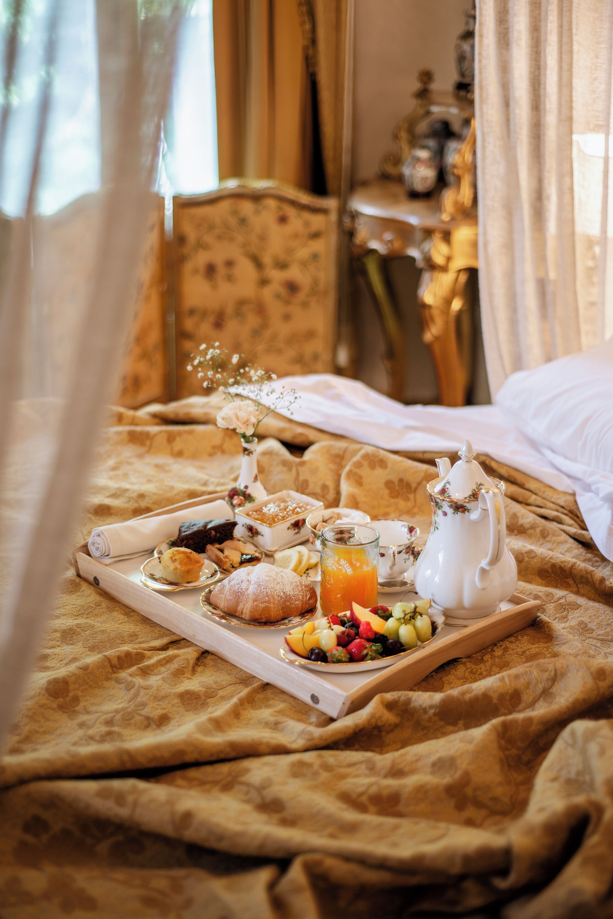 A breakfast tray with pastries, fruits, and a jug of orange juice on a bed with a floral bedspread.