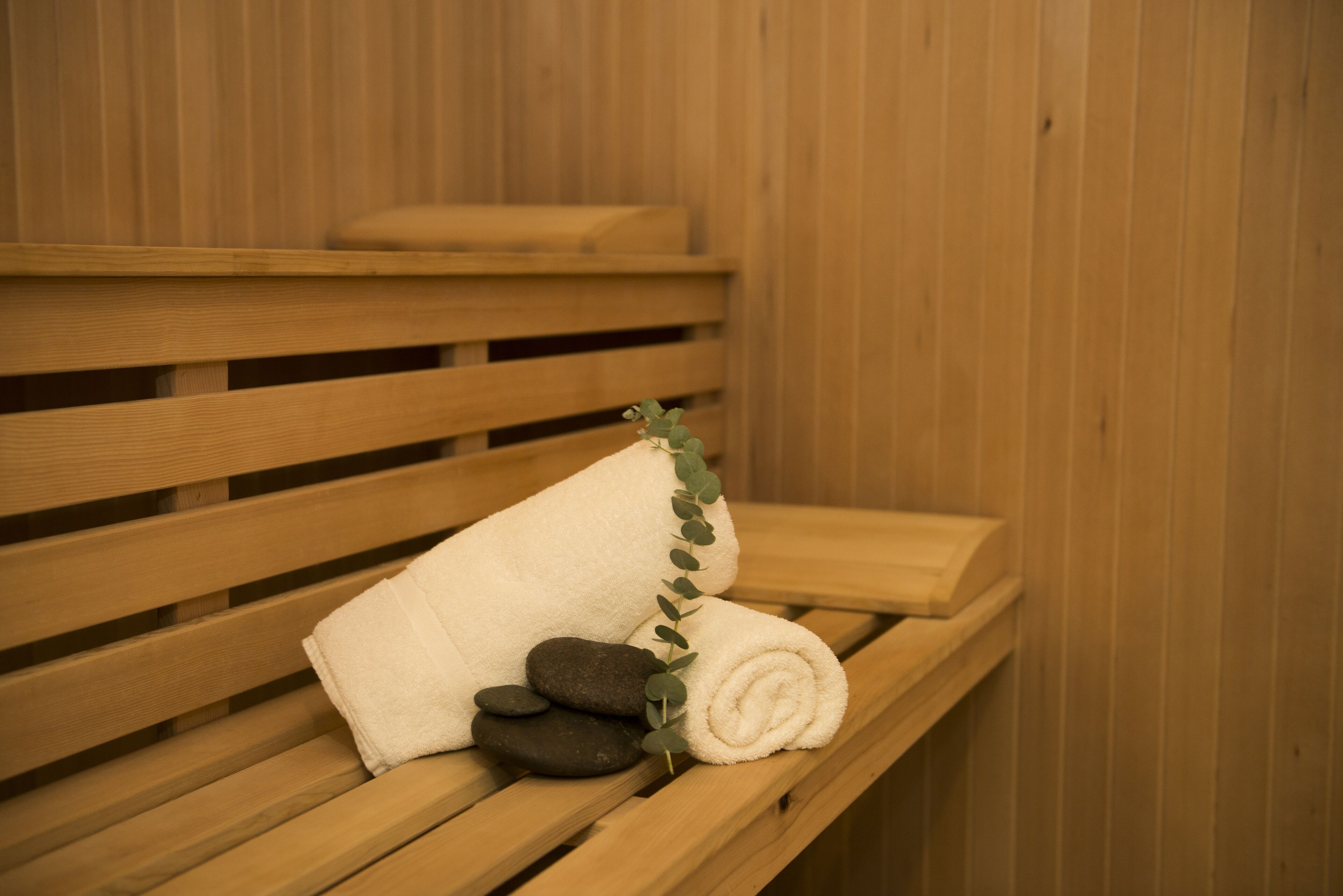 A wooden bench with a rolled white towel and a small pile of dark stones, accompanied by a sprig of greenery.
