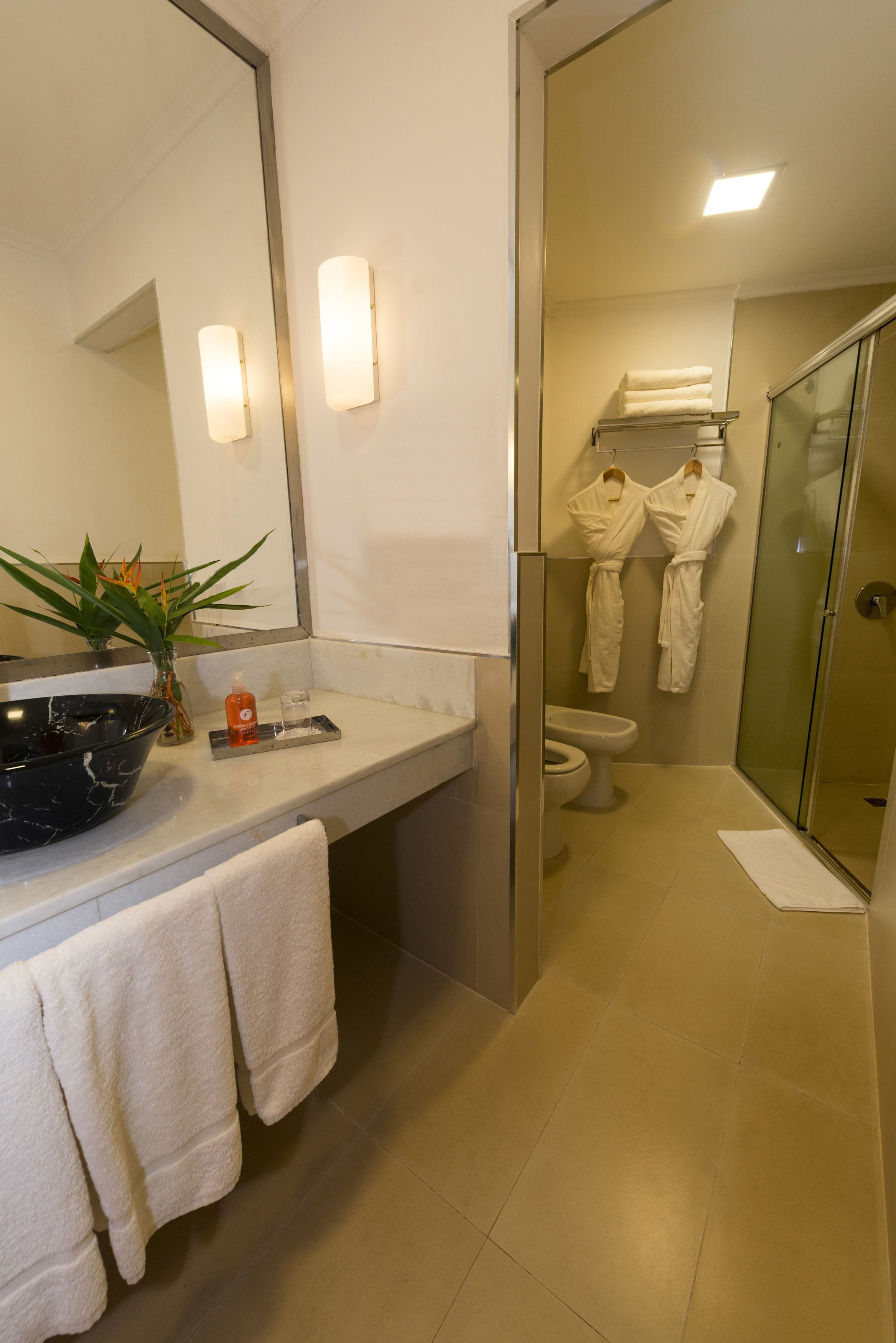 A bathroom with a large mirror, a sink with a vase of plants, a toilet, and a glass-enclosed shower.