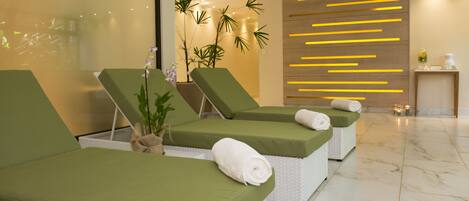 Uma área de spa com três poltronas confortáveis verdes, uma parede com linhas horizontais amarelas e uma pequena mesa com um vaso e uma vela.