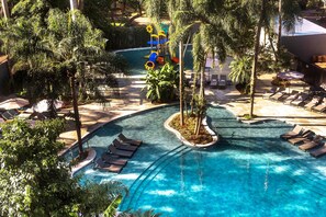 Un espace piscine aménagé dans un complexe hôtelier, avec des chaises longues, des palmiers et un toboggan aquatique.