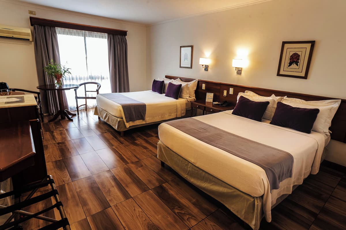 Ein Hotelzimmer mit zwei Betten, einem Schreibtisch, einem Stuhl, einer Zimmerpflanze und einem Vorhangfenster.