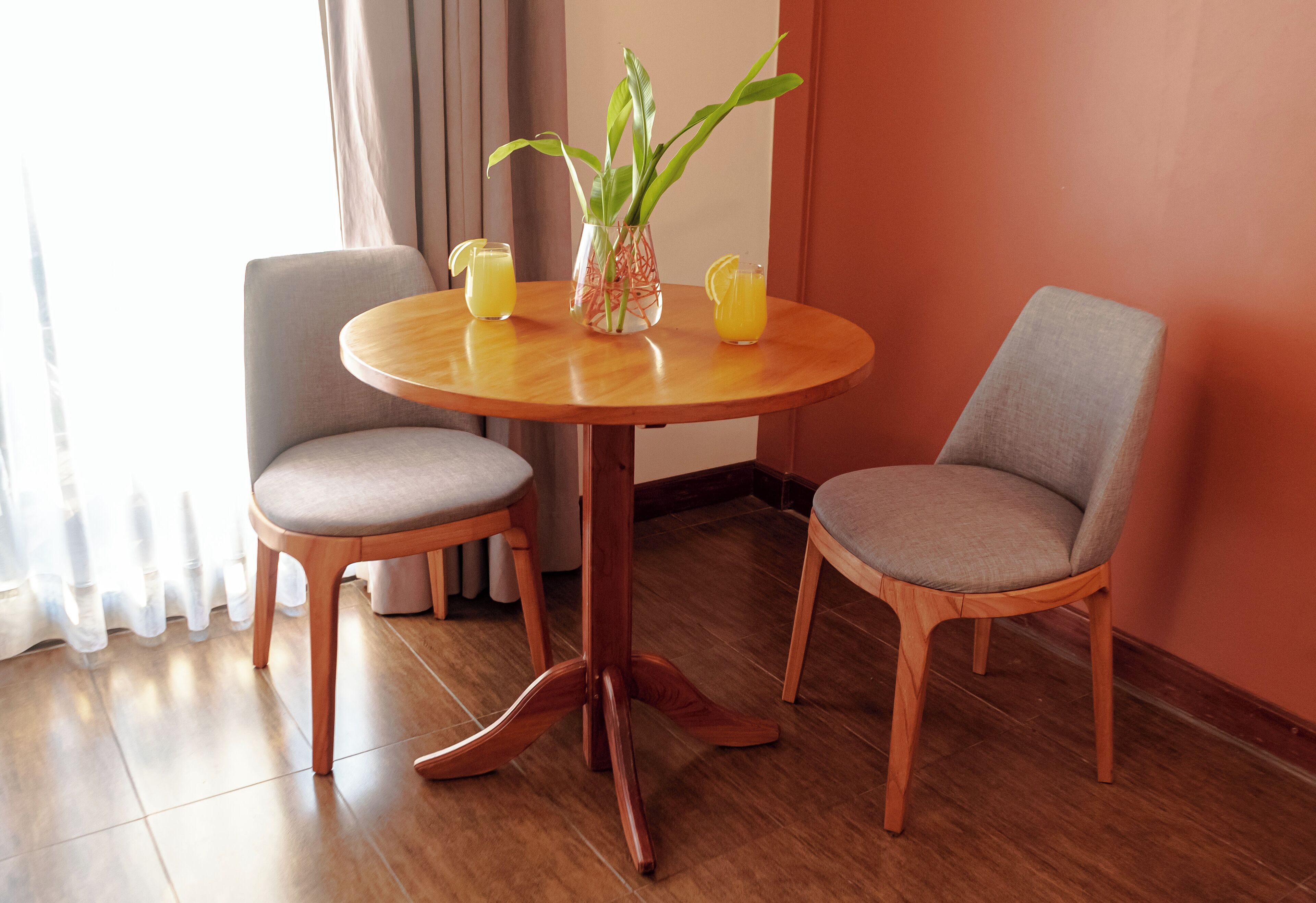 A round wooden table with two grey upholstered chairs, a vase with green plants, and two glasses of orange juice.