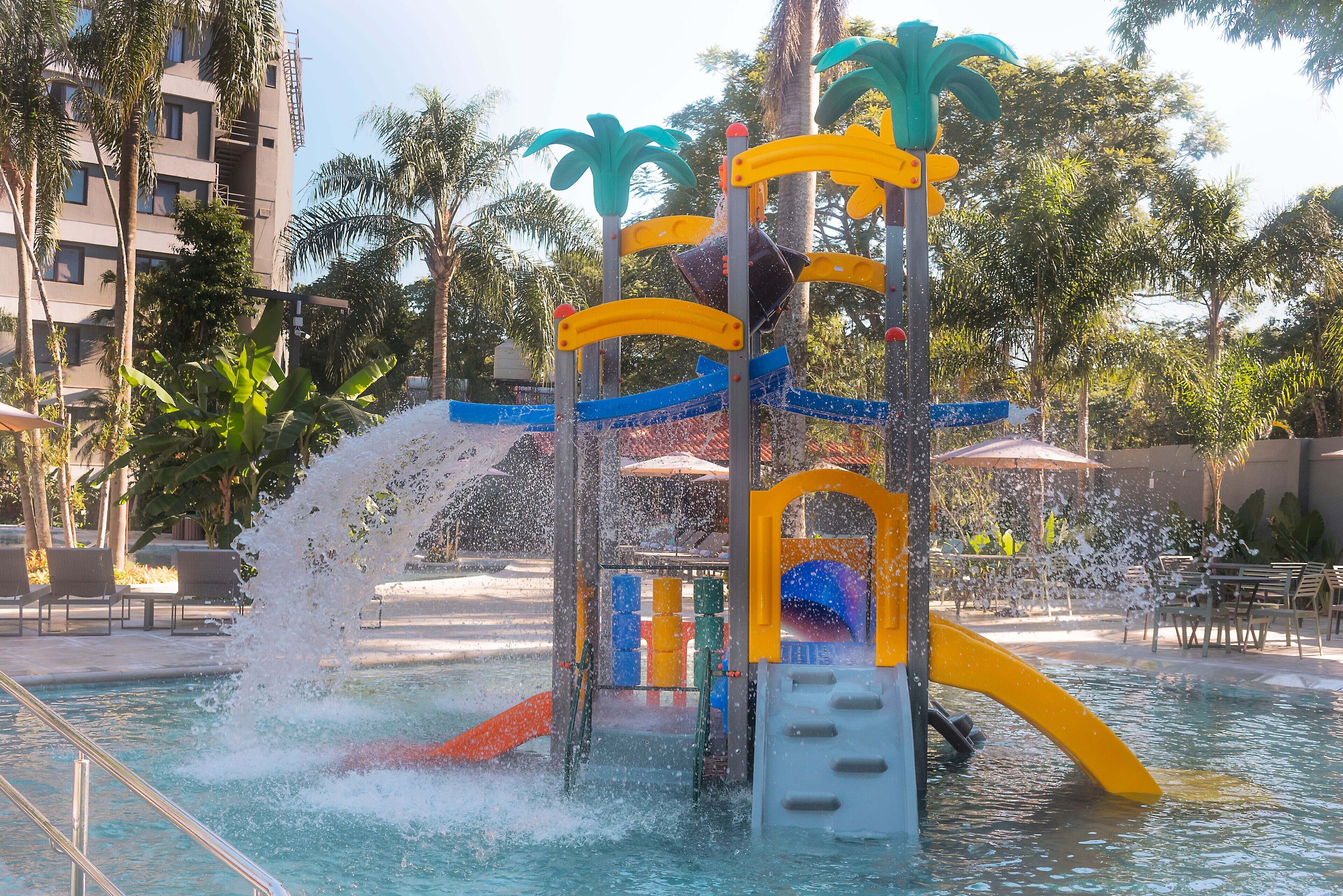 A water play area with slides and water jets, surrounded by palm trees and a pool.
