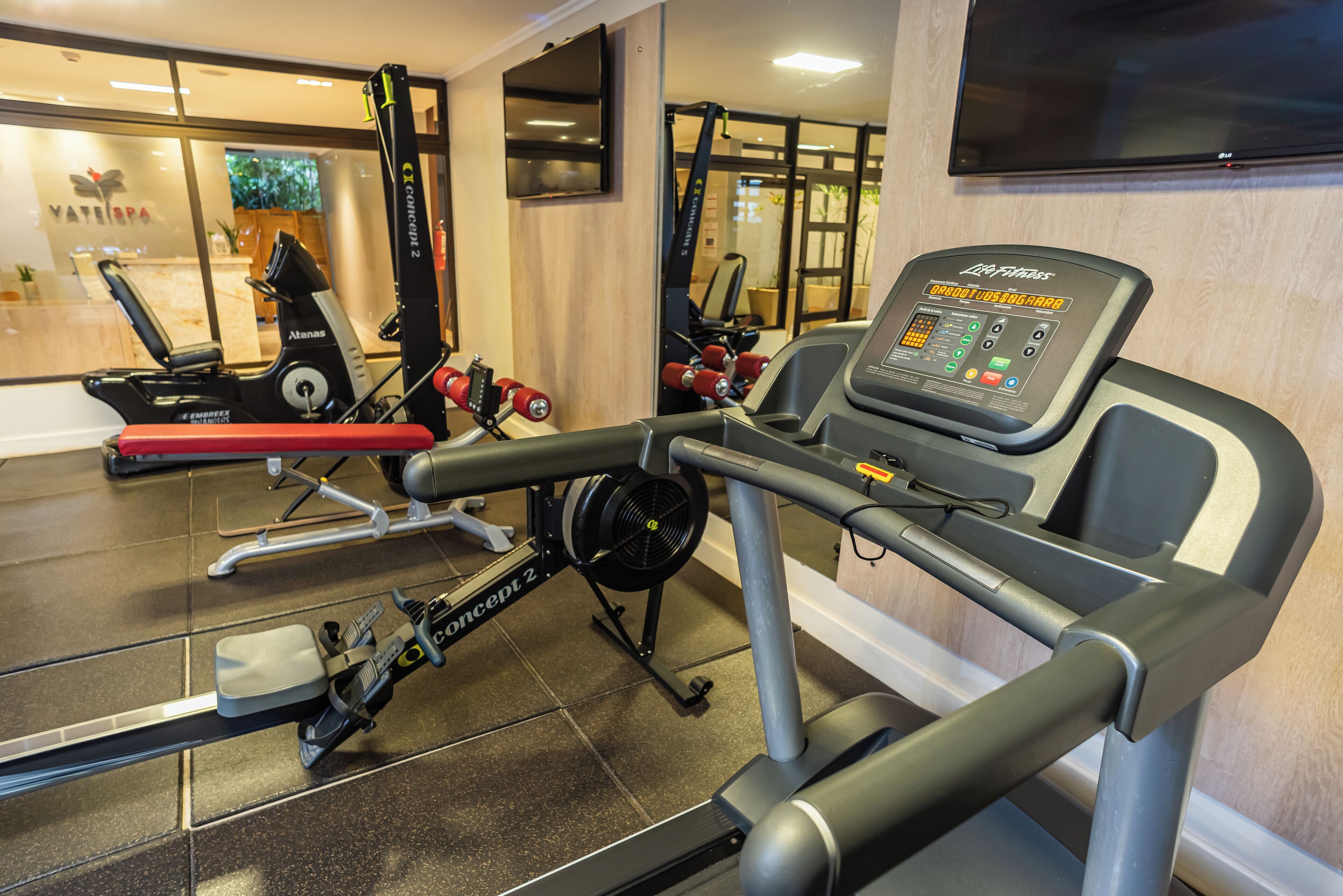 Un gimnasio con máquinas de cardio, equipo de levantamiento de pesas y un televisor en la pared.