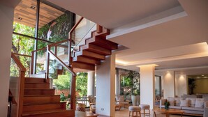 Interior moderno com escada de madeira, sala de estar com sofá e poltronas, e janelas amplas com vista para o verde.