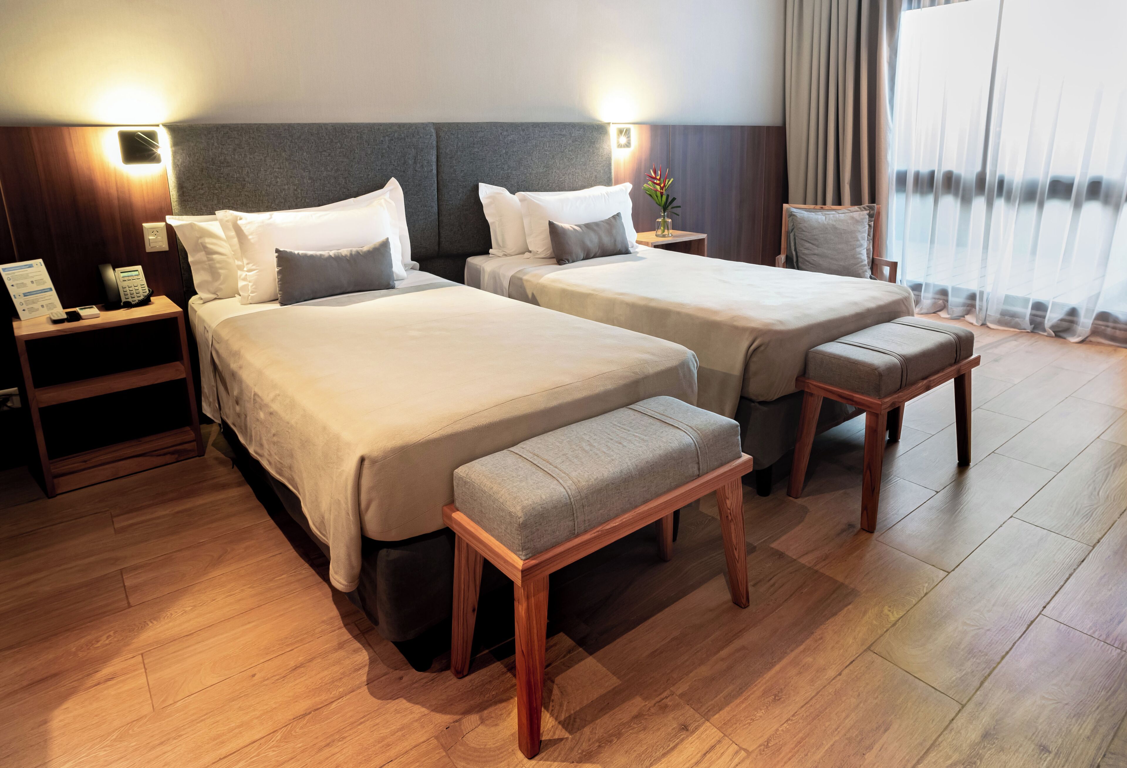 A hotel room with two beds, a wooden bench, a nightstand with a phone, and a view of the outdoors through sheer curtains.