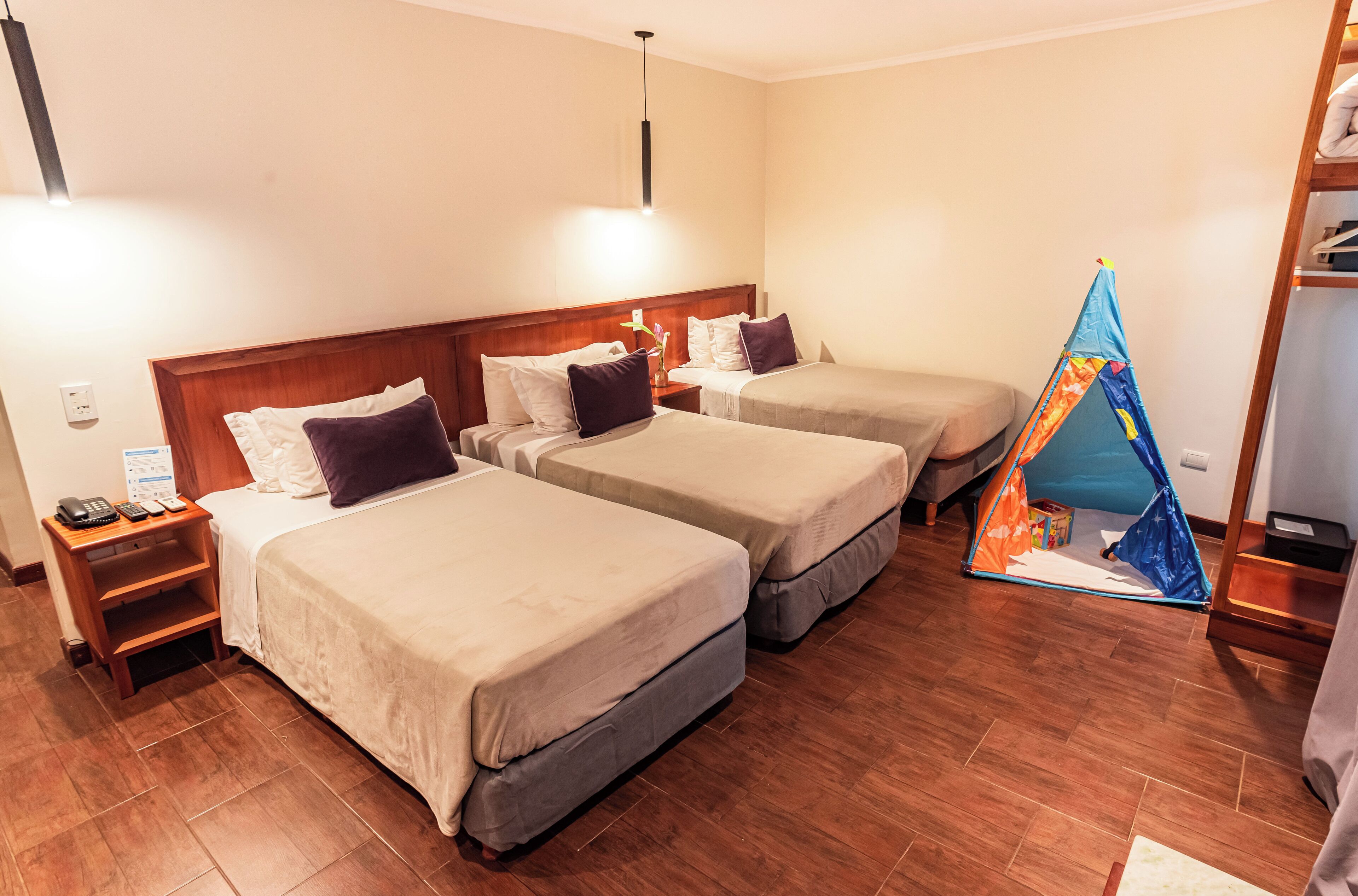 A hotel room with three single beds, wooden headboards, a small table with a vase, and a tent-like structure.