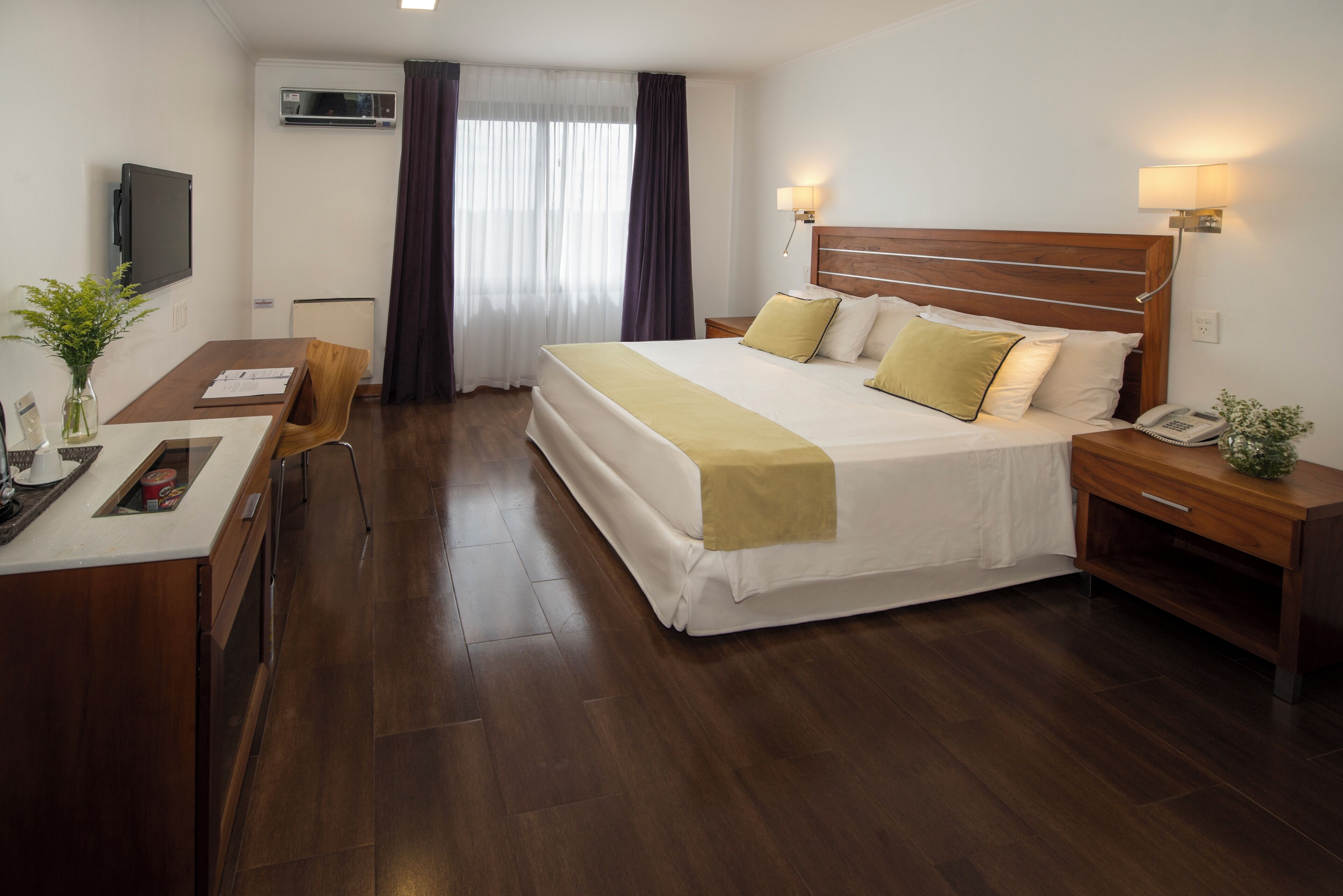 A hotel room with a large bed, bedside tables, a desk with a chair, a TV, and a window with curtains.