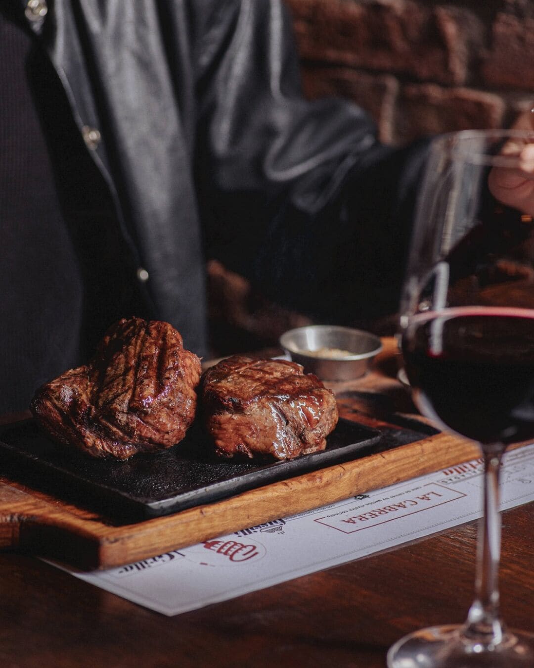 A steak on a wooden board, a glass of red wine, and a menu on a table.