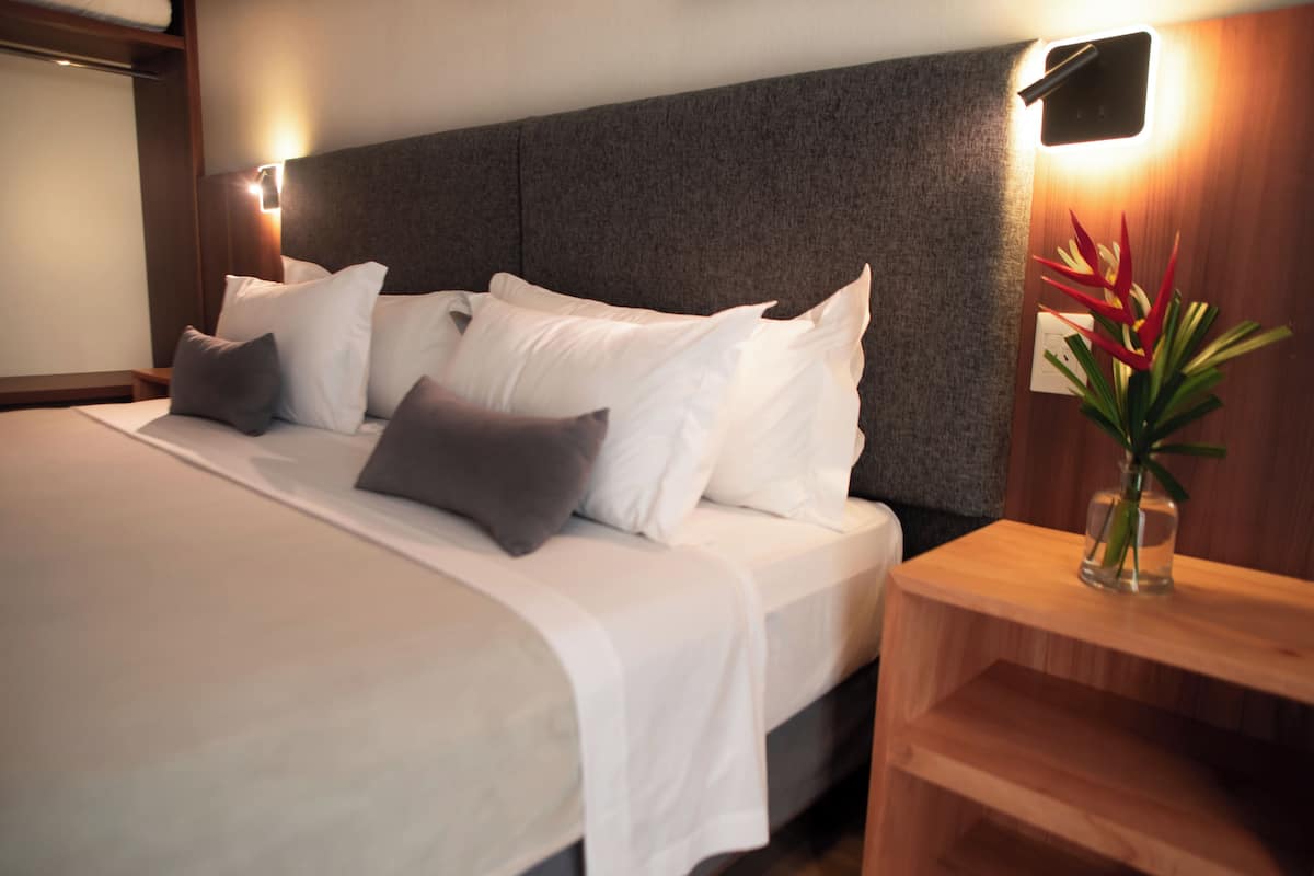 Ein ordentlich bezogenes Bett mit weißer und grauer Bettwäsche, ein Holz-Kopfteil und ein Nachttisch mit Blumen in einer Vase.