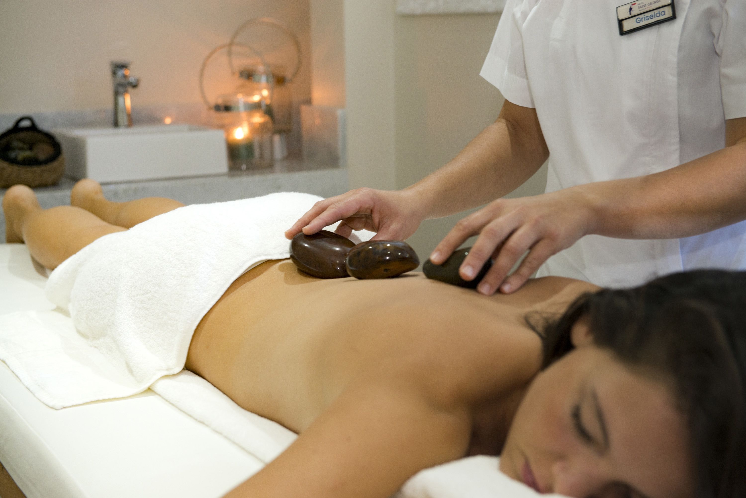 A massage therapy session in a spa setting with a client lying on a treatment table, a therapist using stones on the client's back, and a sink and candle in the background.