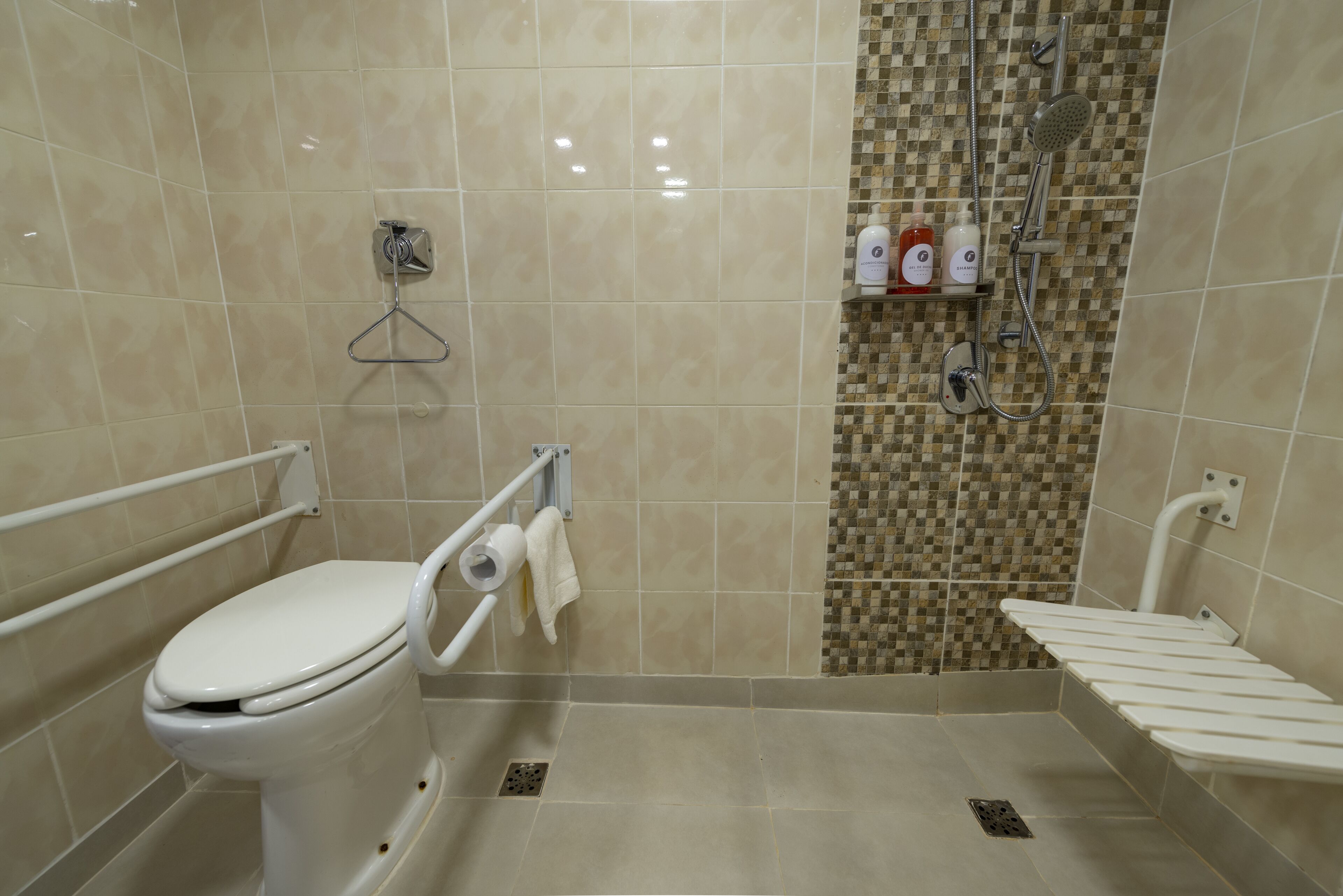 Un baño con inodoro, barras de apoyo, zona de ducha con pared de azulejos y estante con artículos de aseo.
