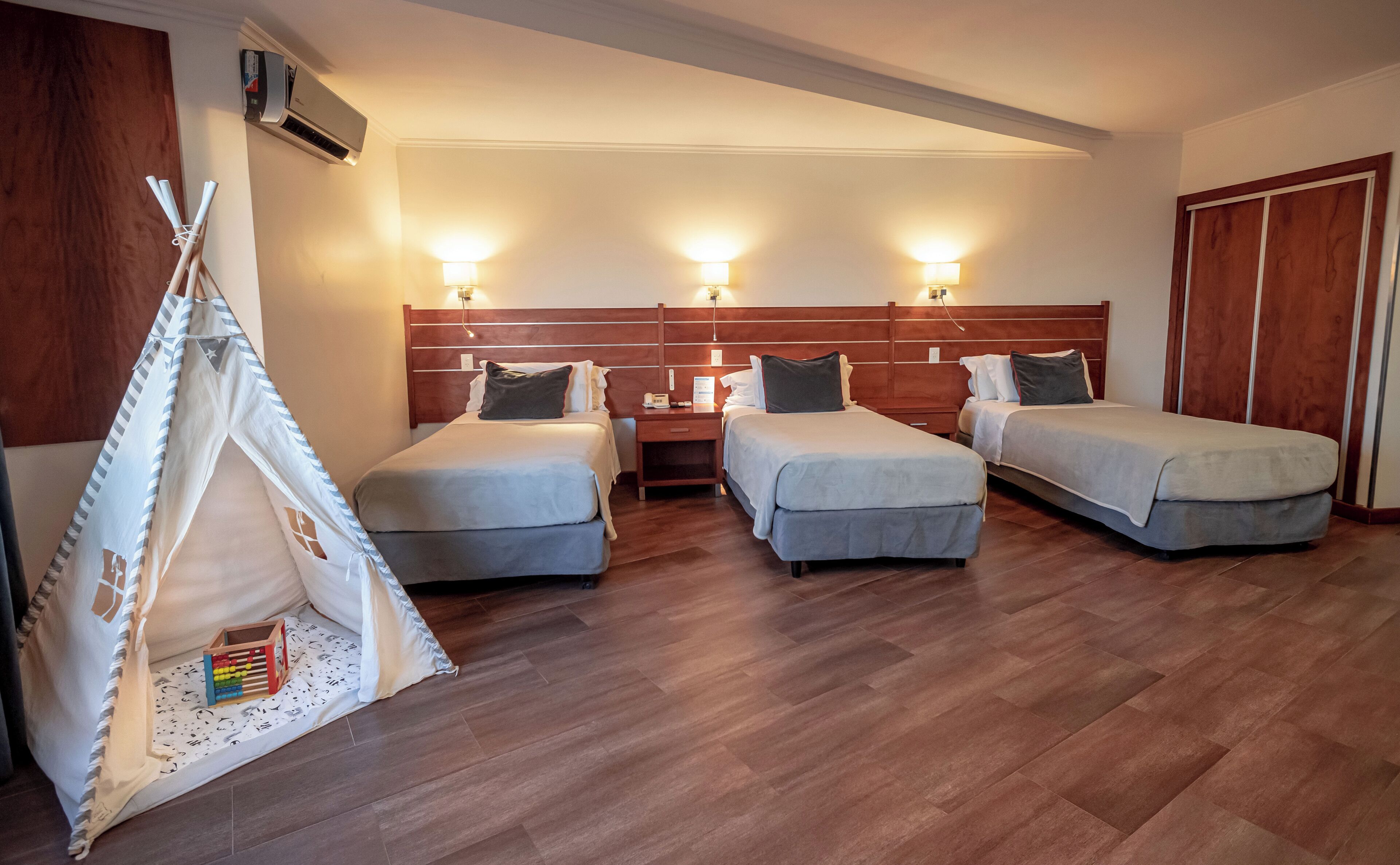 A hotel room with three single beds, a wooden headboard, a nightstand, a lamp, and a wooden wardrobe.