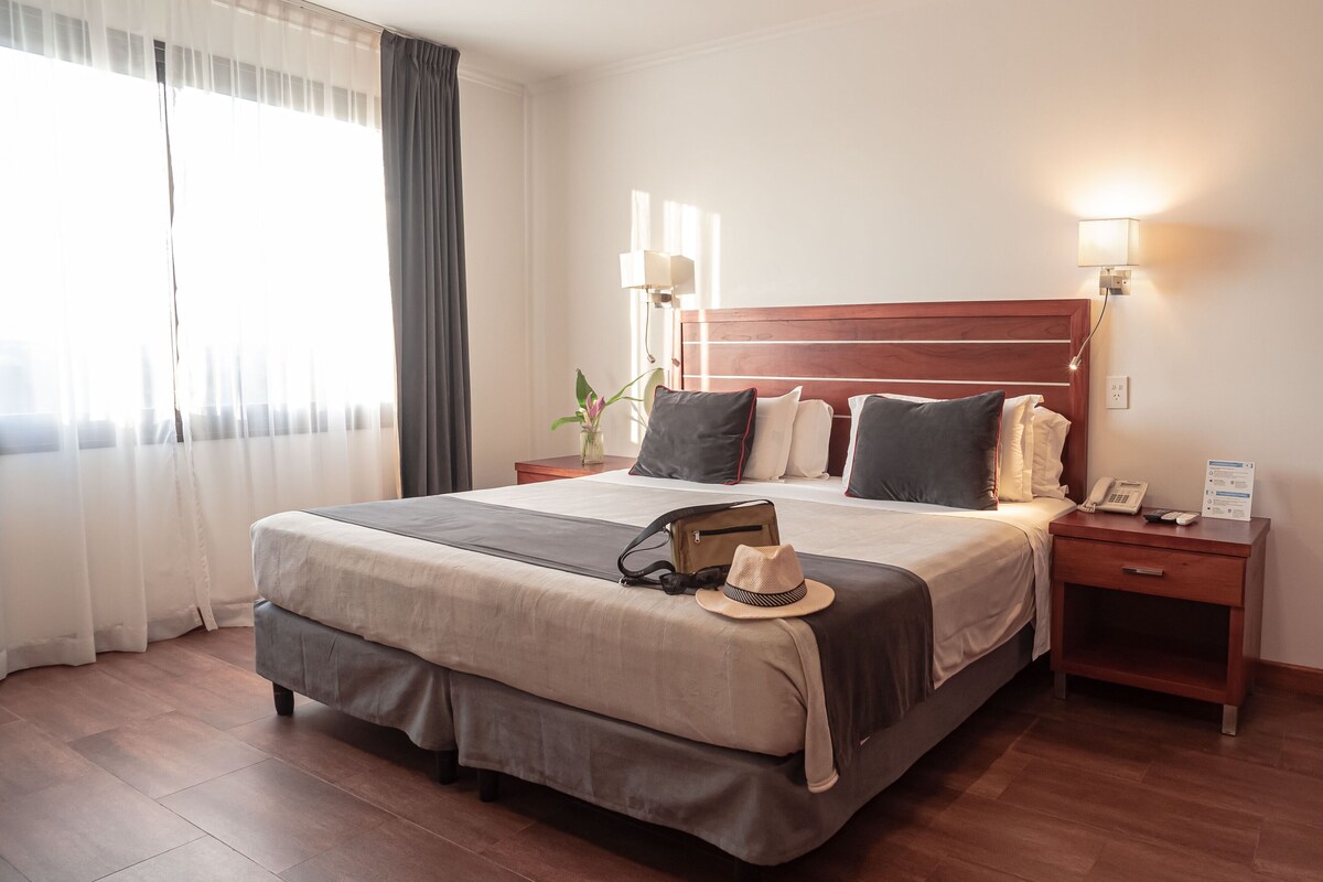 Een hotelkamer met een groot bed, bedlampjes, een nachtkastje met telefoon en een raam met gordijnen.