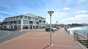 A coastal walkway with a modern building, parked cars, and a view of the sea. - Hotel St Clair (Dunedin)