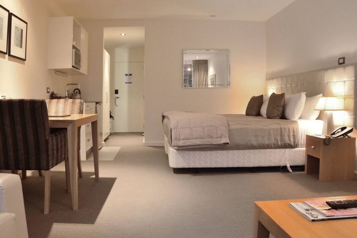 A hotel room with a bed, bedside table, lamp, and a desk with a chair.