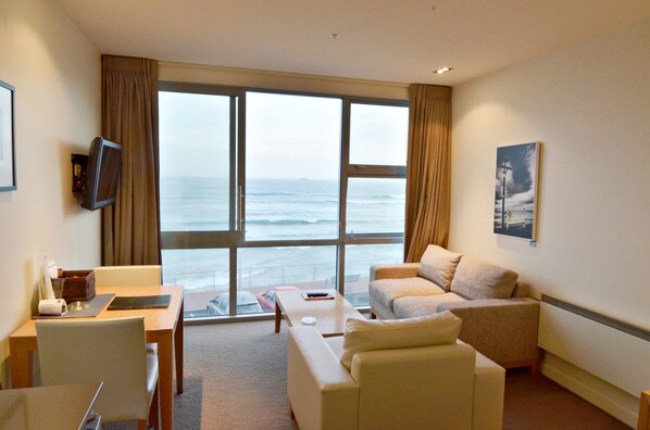 A hotel room with a large window offering a view of the sea, a flat-screen TV mounted on the wall, a sofa, a small table, and a chair.