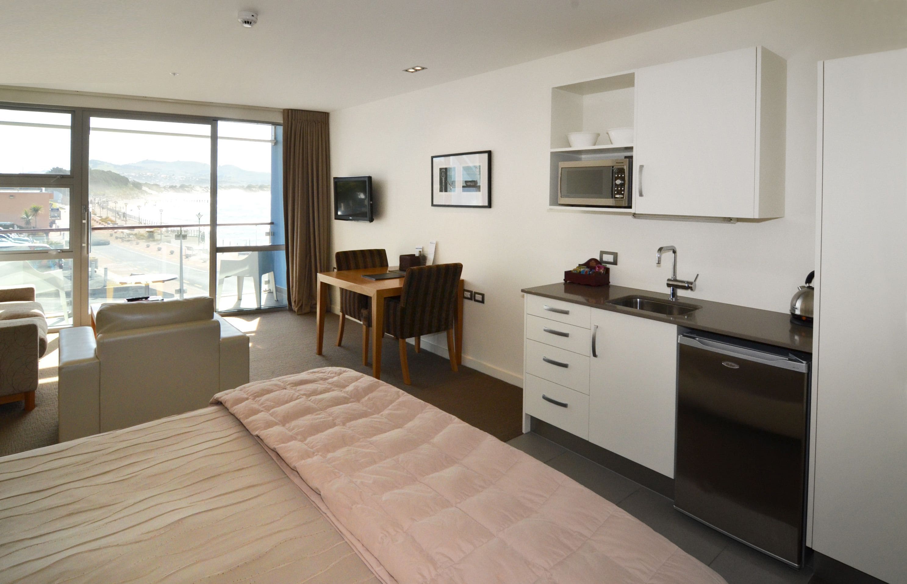 A hotel room with a bed, a desk, a chair, a small kitchenette, and a view of the beach.