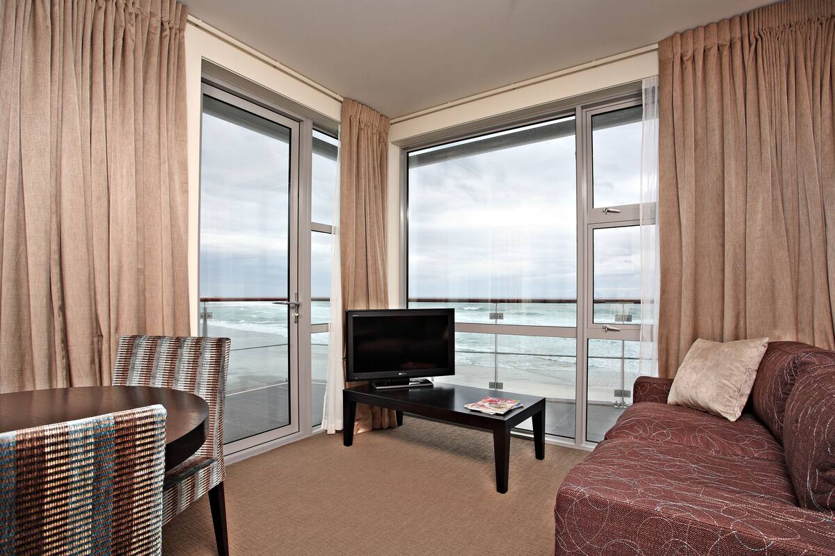 A room with a TV on a table, a sofa, a chair, and large windows overlooking the sea.