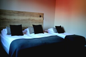 Una habitación de hotel con cabecera de madera, cama con ropa de cama blanca y varios almohadones, y una manta oscura.