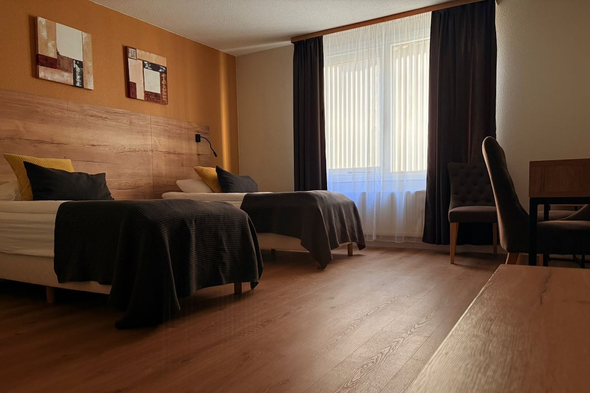 Habitación de hotel con dos camas, cabecera de madera, escritorio, silla y ventana con cortinas.