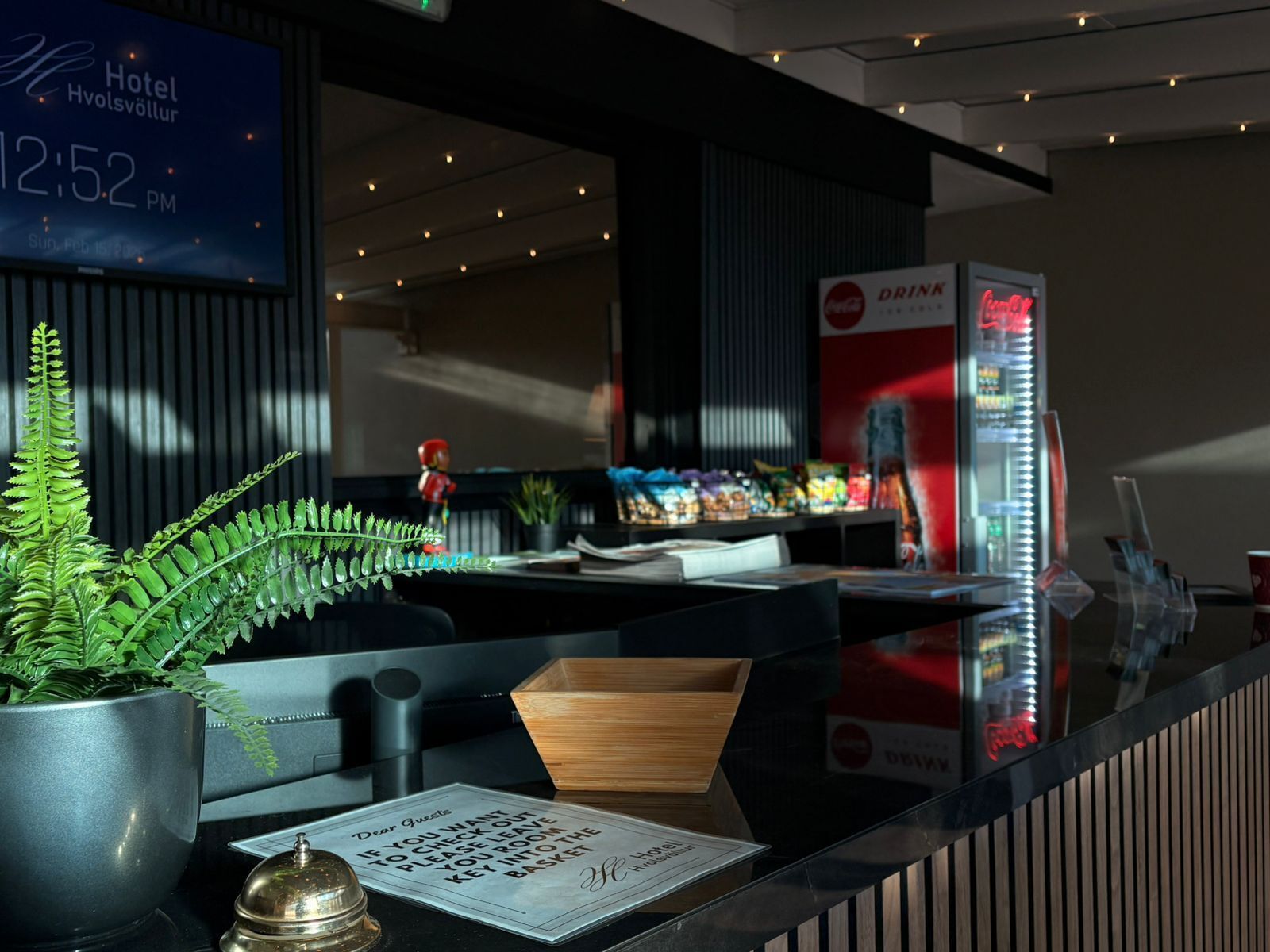 A hotel reception desk with a digital clock displaying 12:52 PM, a Coca-Cola vending machine, a fern plant, and a wooden box.