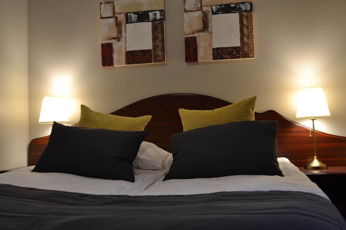 Una cama bien tendida con dos almohadas, cabecera de madera y dos lámparas fijadas a la pared.