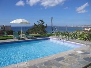 A swimming pool with a view of the sea, surrounded by stone tiles and two lounge chairs under a white umbrella.