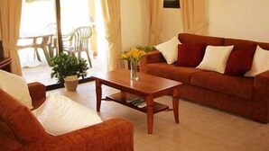 A living room with a sofa, a coffee table, and a chair. There is a window with curtains, a plant on the table, and a view of the outside. - Aura Holiday Villas (Kissonerga)