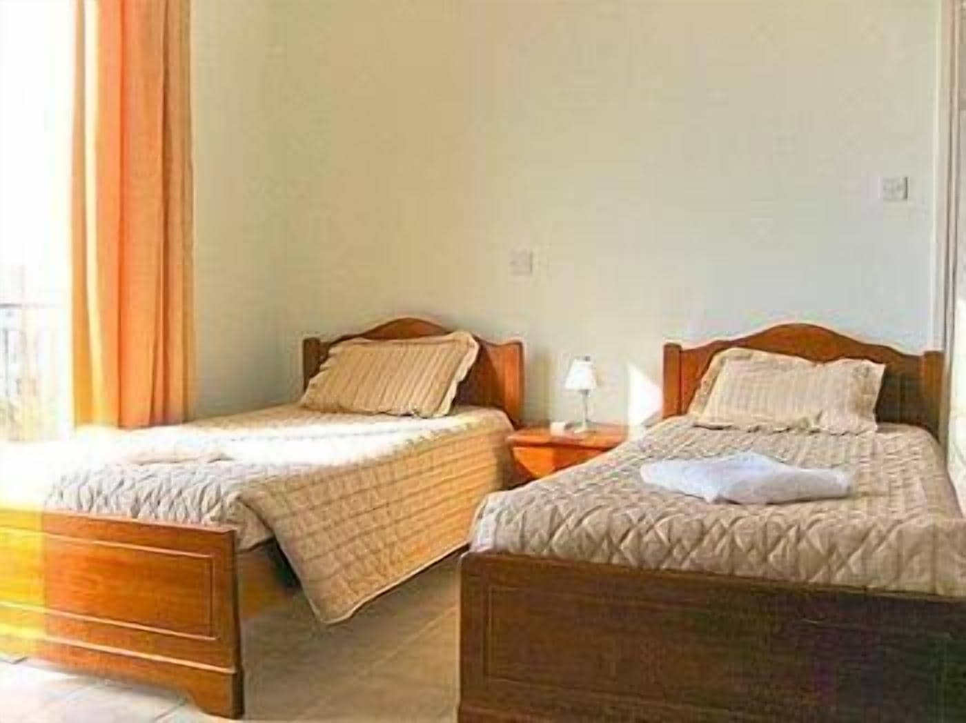 A hotel room with two single beds, wooden headboards, a nightstand with a lamp, and a window with curtains.