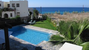 A swimming pool with a view of the ocean, surrounded by a white building with a balcony and a garden area with a palm tree. - Aura Holiday Villas (Kissonerga)