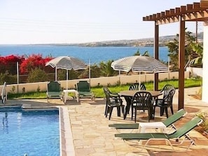 A pool area with lounge chairs and umbrellas, overlooking a body of water. - Aura Holiday Villas (Kissonerga)