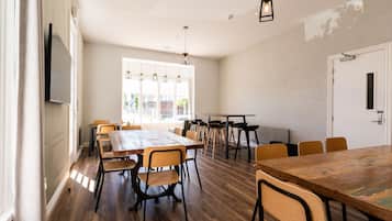 A spacious room with a large wooden table and several chairs, a flat-screen TV mounted on the wall, and a large window providing natural light.