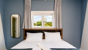 Una cama bien tendida con sábanas blancas y una toalla enrollada, una ventana con cortinas y un espejo en la pared.
