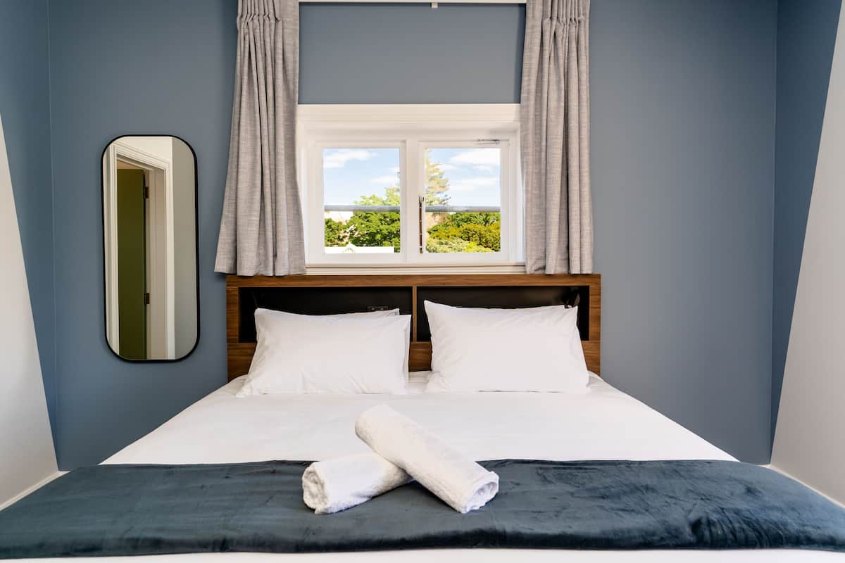 Una cama bien tendida con sábanas blancas y una toalla enrollada, una ventana con cortinas y un espejo en la pared.