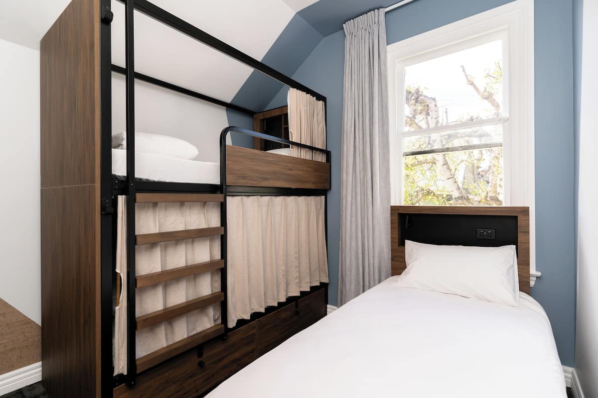Una cama litera con estructura de madera, una cama inferior con colchón blanco y una ventana con cortinas.