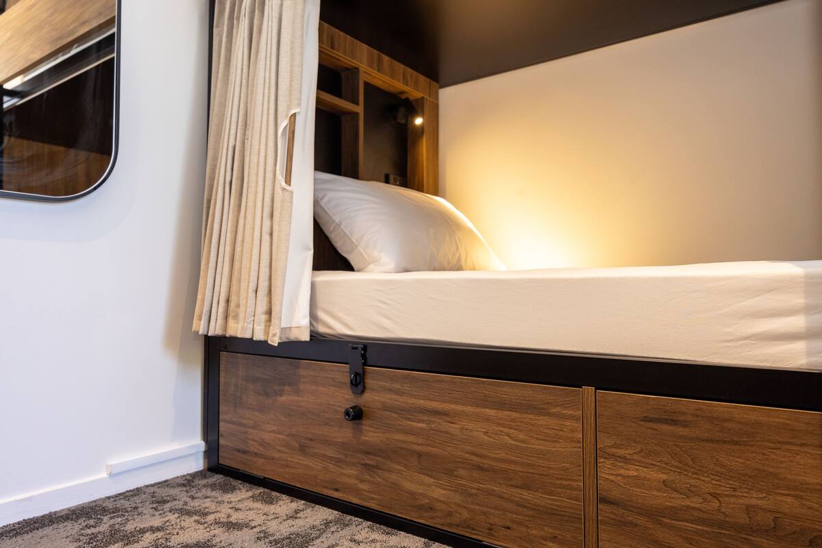 Una cama compacta con estructura de madera y colchón blanco, una mesita de noche pequeña y una cortina.