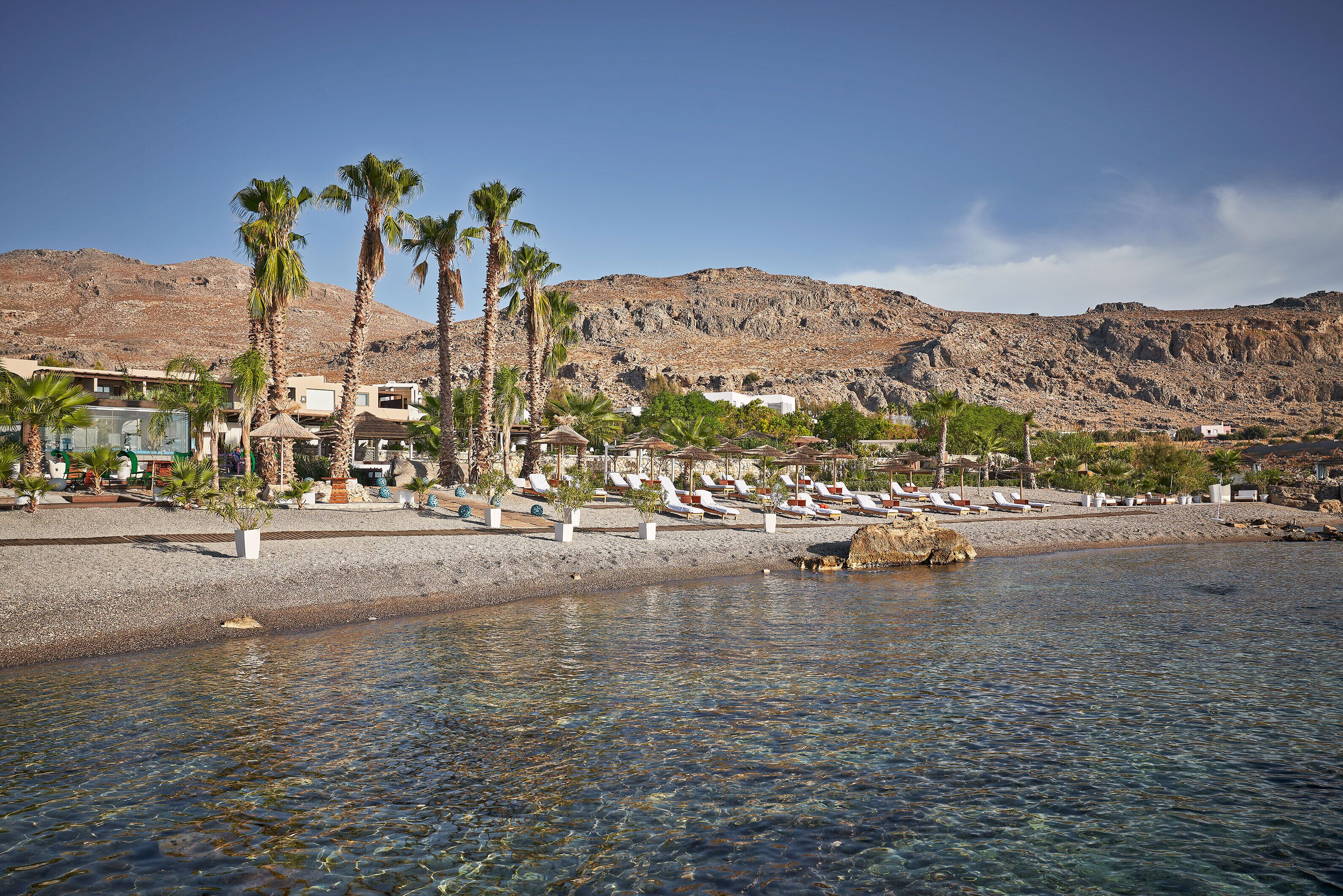 A beach resort with palm trees, clear water, and a mountainous backdrop.