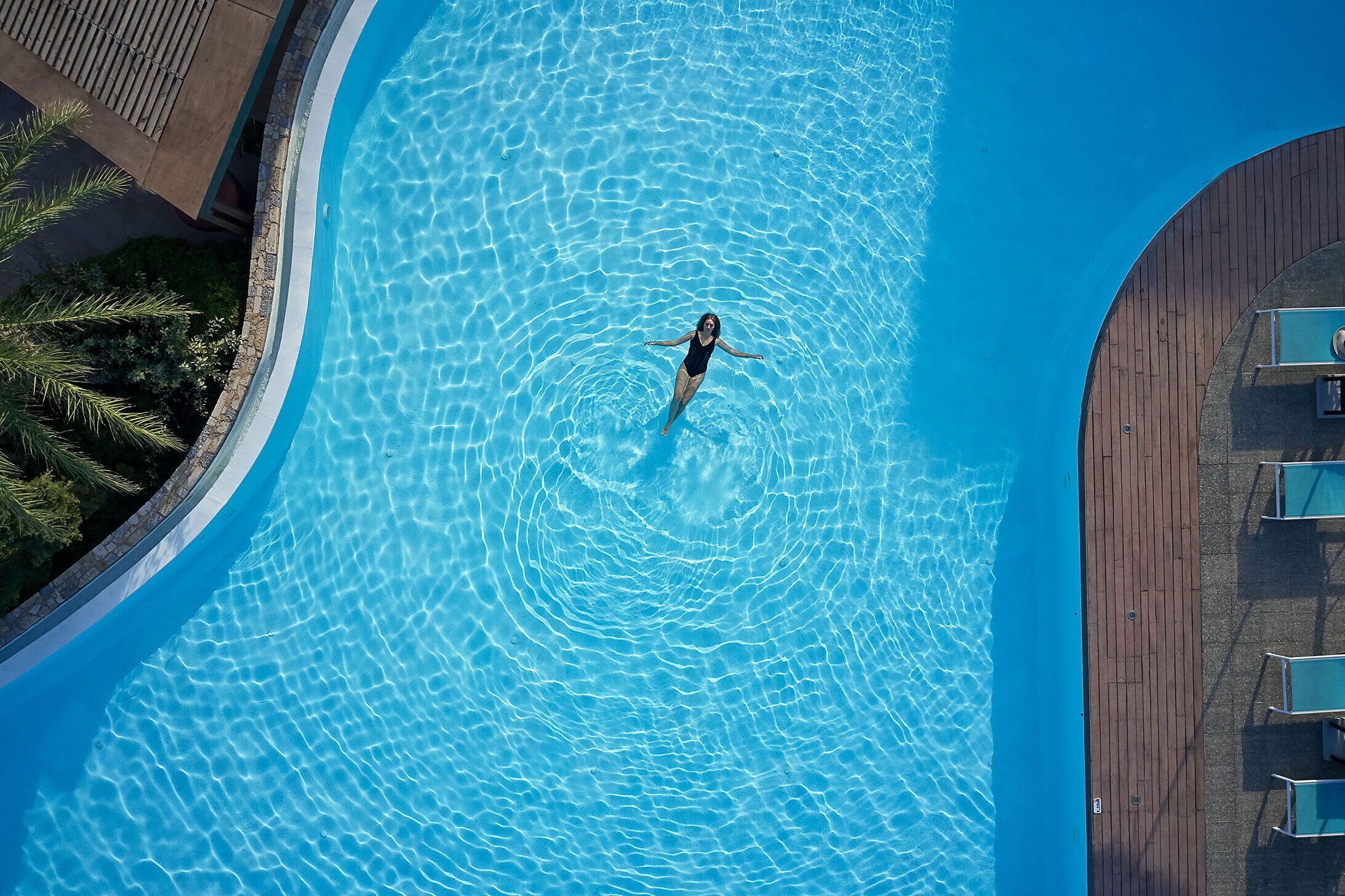 Vista aérea de una piscina con una persona flotando en el centro, rodeada por una terraza de madera y sillones de descanso.
