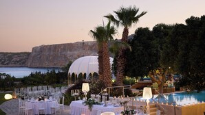 Area pranzo all'aperto con tavoli apparecchiati per un evento formale, con vista su una piscina e una scogliera sul mare.