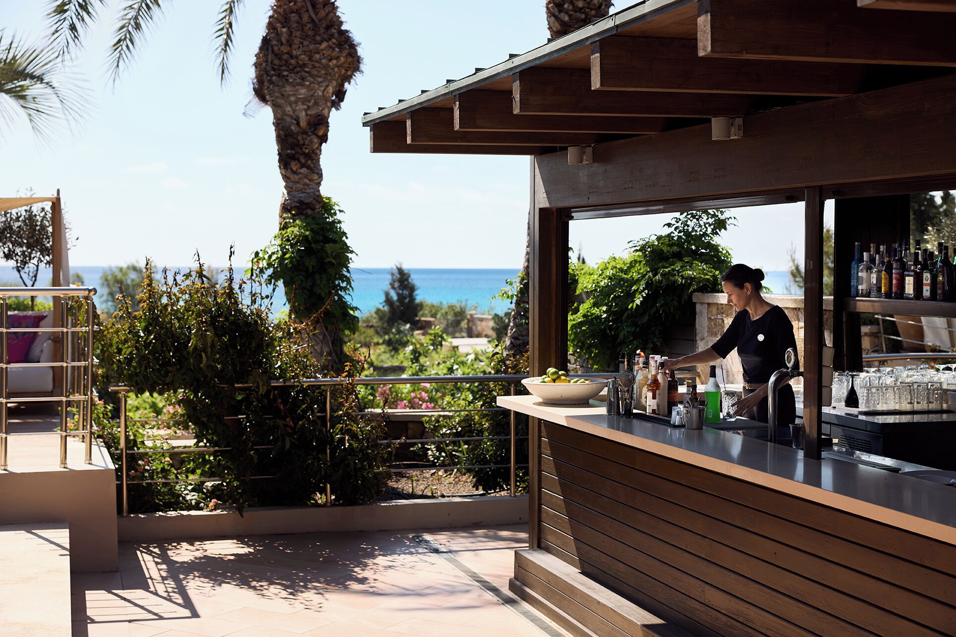 A bar area with a view of the sea, a bartender preparing drinks, and a seating area with a railing.