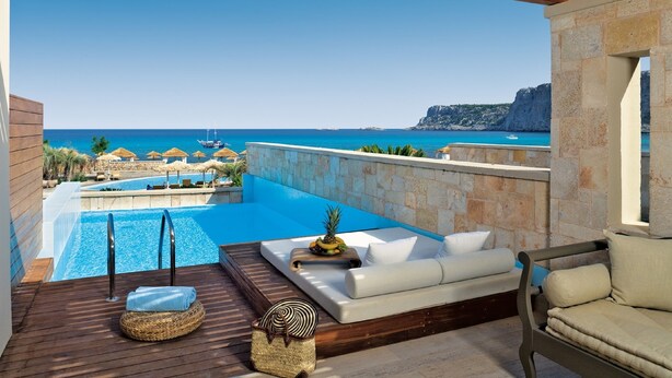 Area piscina con vista sul mare, un pontile in legno e un angolo relax con cuscini e un tavolino con frutta.
