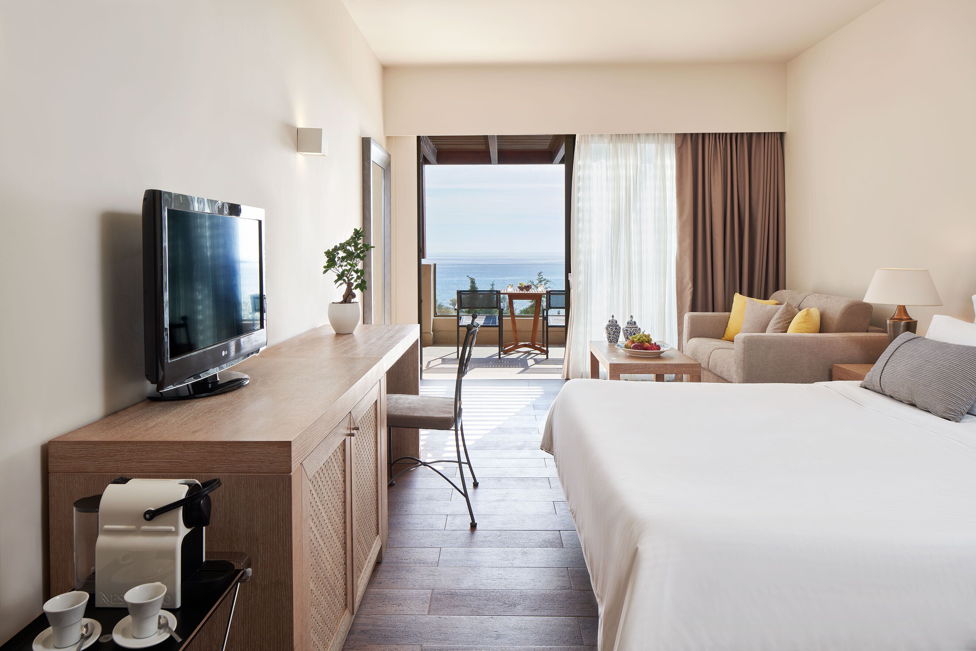 Habitación de hotel con cama, televisión, escritorio con cafetera, silla y balcón con vista al océano.