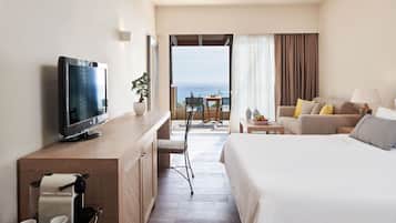 Habitación de hotel con cama, televisión, escritorio con cafetera, silla y balcón con vista al océano.