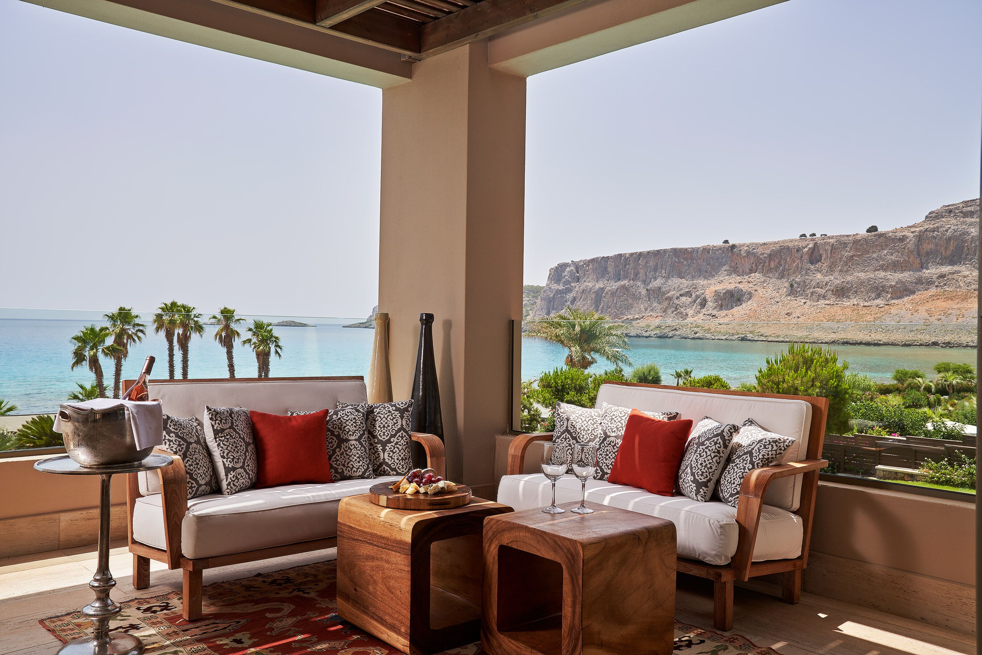 A balcony with a sofa and chairs, a small table with a tray of fruit, and a view of the sea and cliffs.